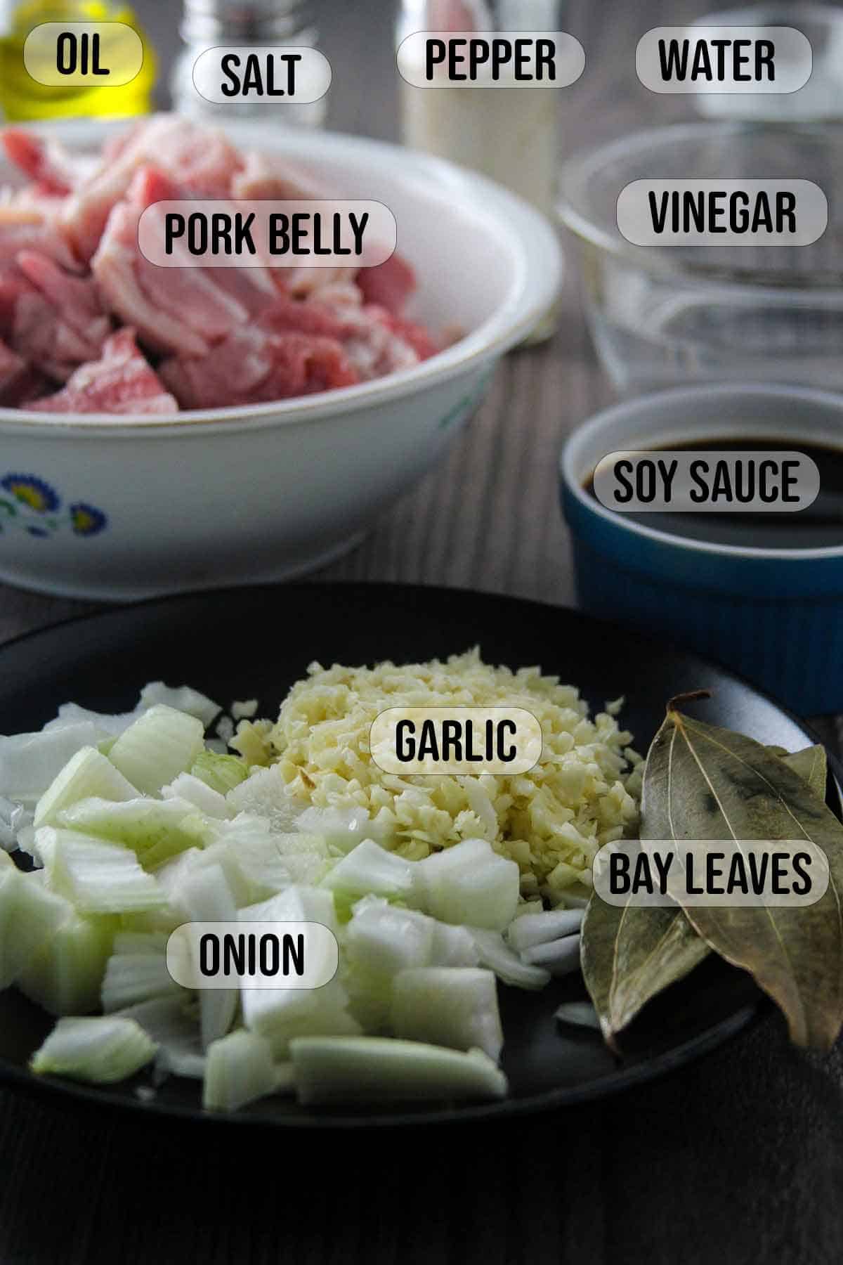 chopped onions, minced garlic, bay leaves on a black plate, soy sauce in a small blue bowl, vinegar in a clear bowl, and pork belly cubes in a white bowl in the background.