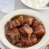 pork adobo in a white bowl with plate of steamed rice and glass of water in the background.