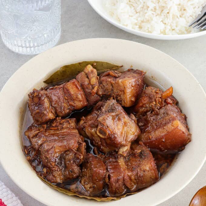 pork adobo in a white bowl with plate of steamed rice and glass of water in the background.