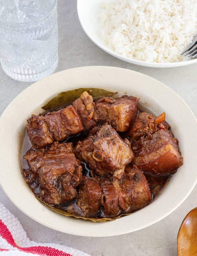 pork adobo in a white bowl with plate of steamed rice and glass of water in the background.