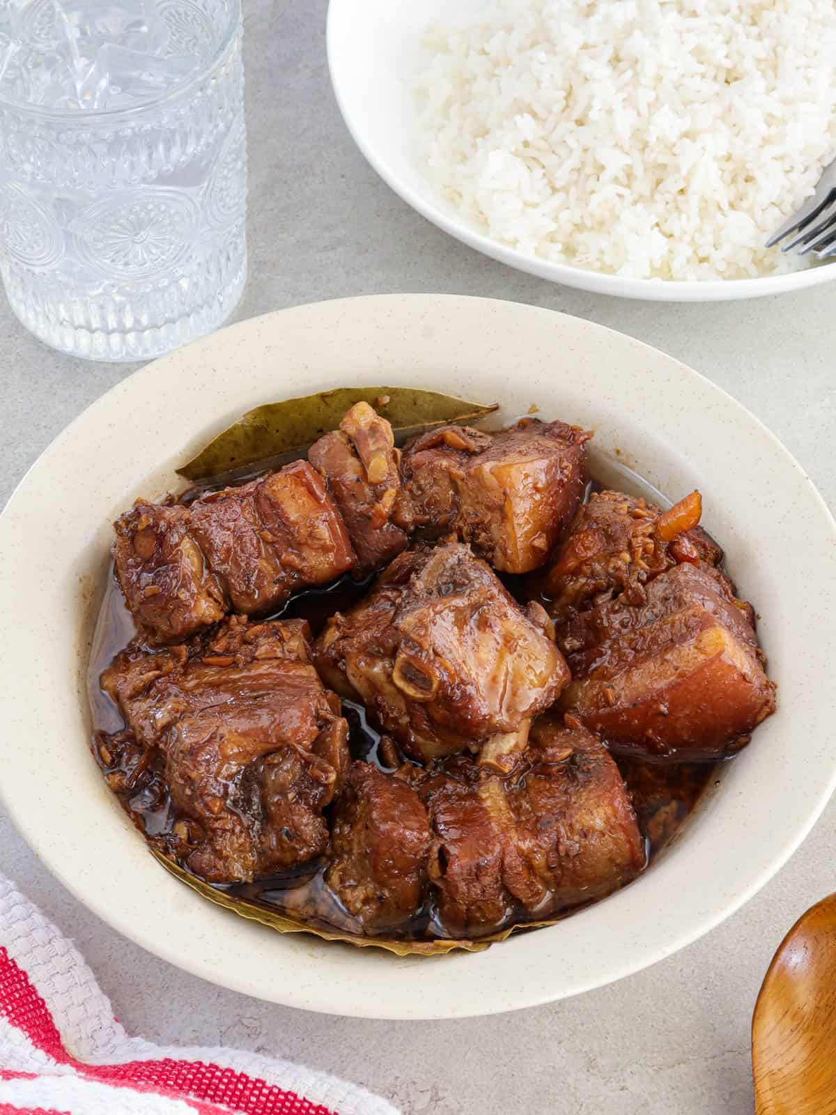 pork adobo in a white bowl with plate of steamed rice and glass of water in the background.