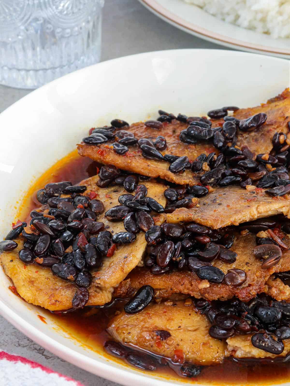 Tilapia fillets in a spicy tausi and garlic sauce on a white serving platter.