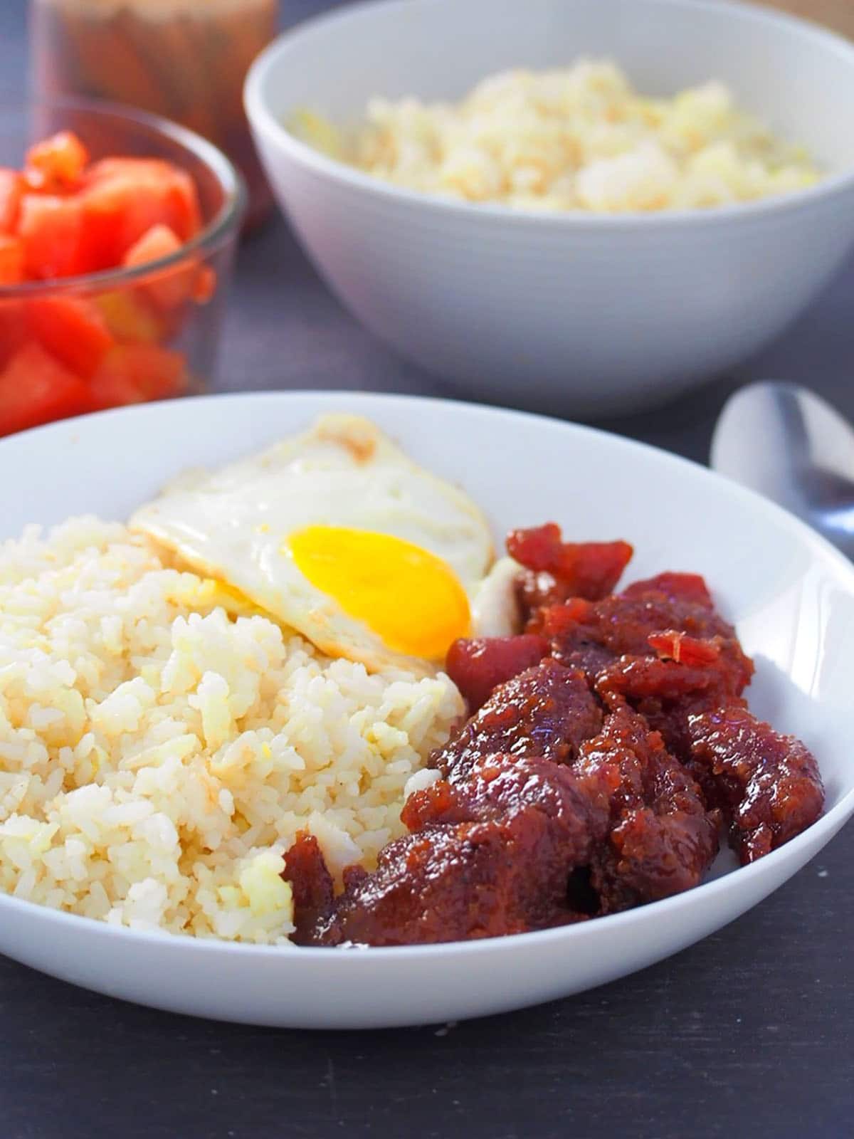 pork tocino with fried rice and fried egg on a white serving palte with a bowl of diced tomatoes on the side