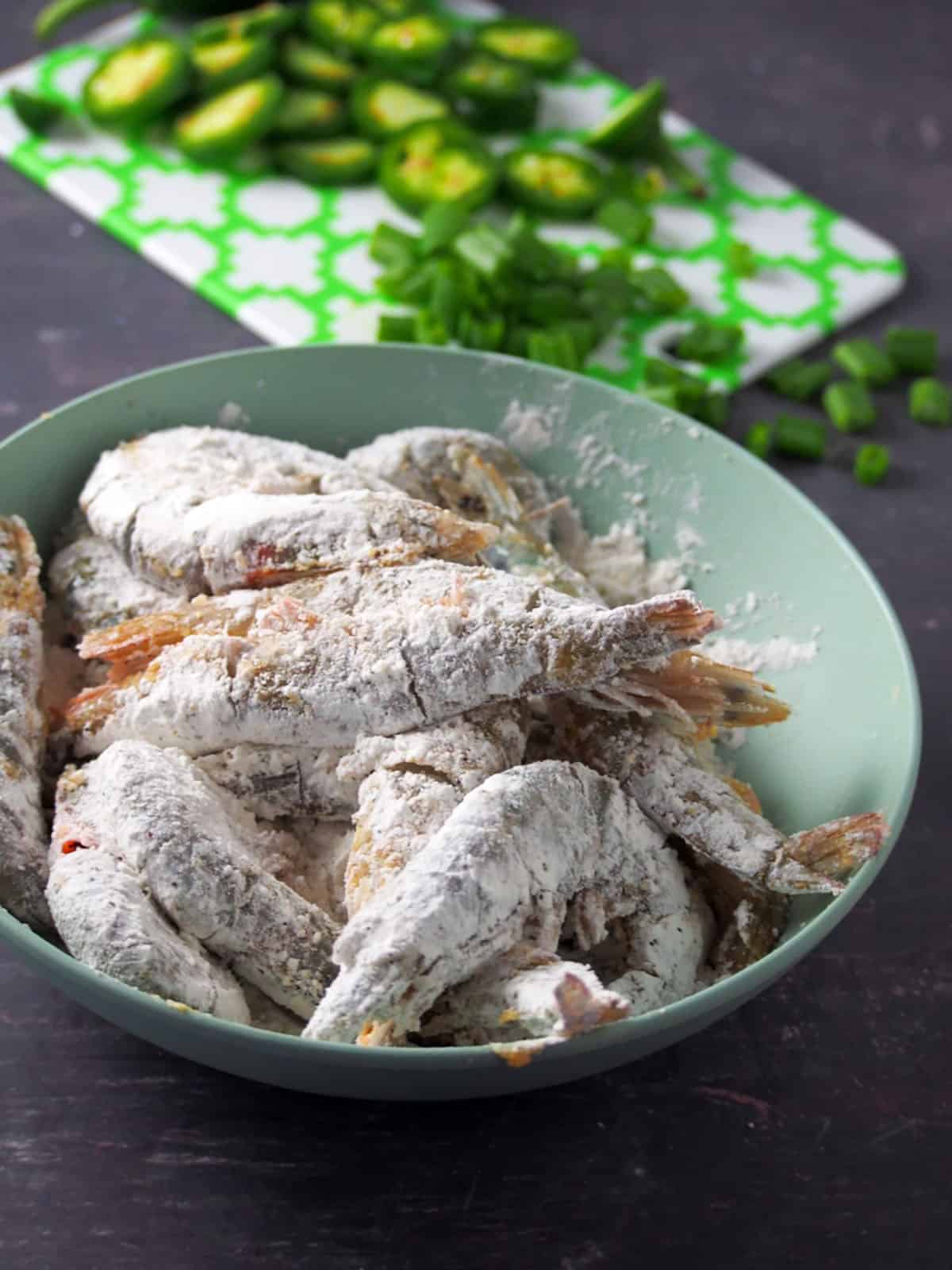 shrimp coated in flour in a blue bowl and sliced jalapeno peppers and chopped green onions on a green chopping board in the background.