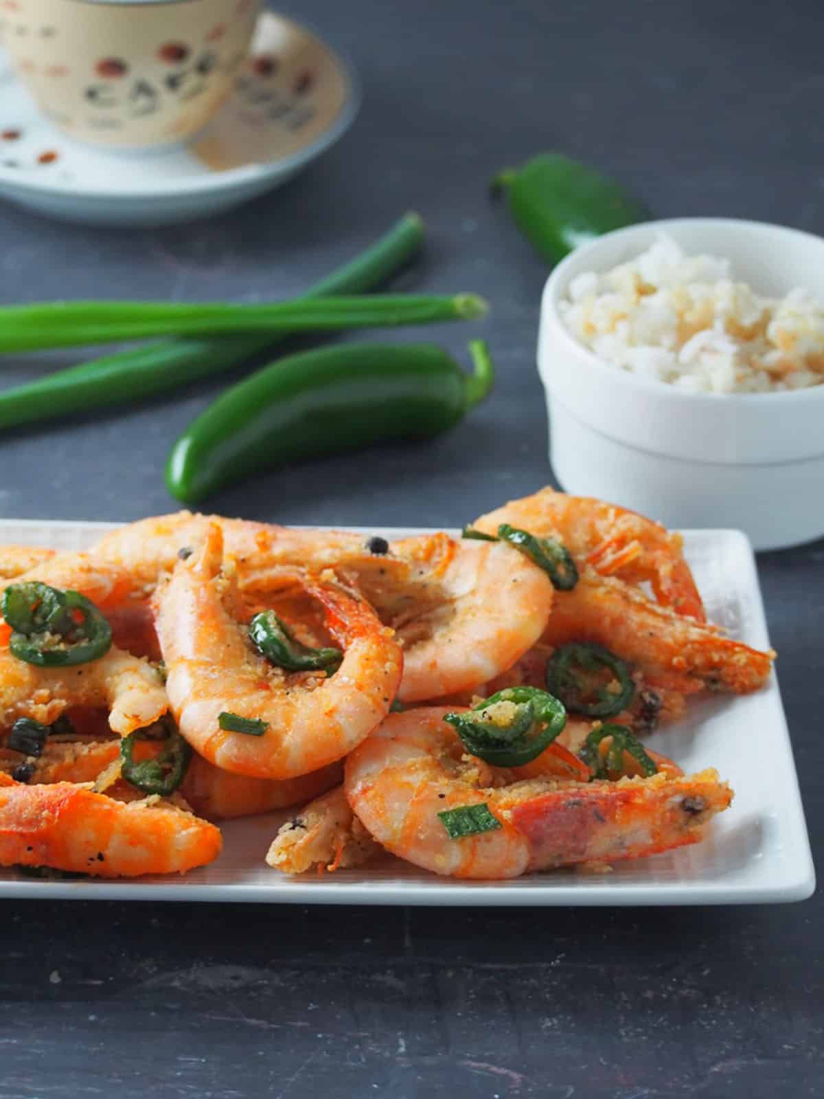 Salt and Pepper Shrimp on a serving platter with jalapeno, green onions, and a bowl of rice in the background.