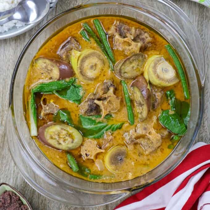 kare-kare in a glass serving bowl with a plate of steamed rice in the background.