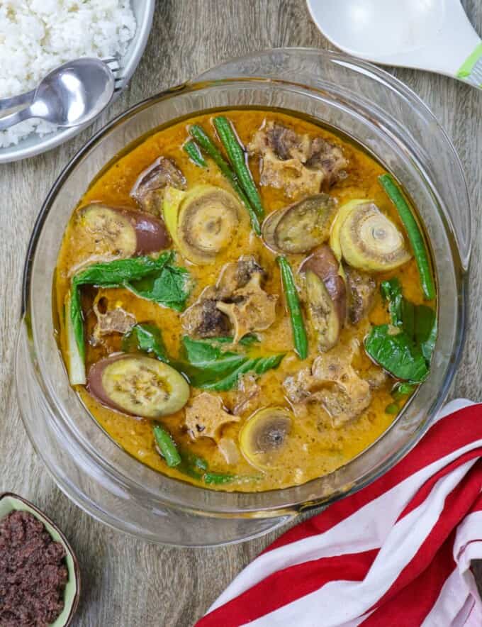 kare-kare in a glass serving bowl with a plate of steamed rice in the background.