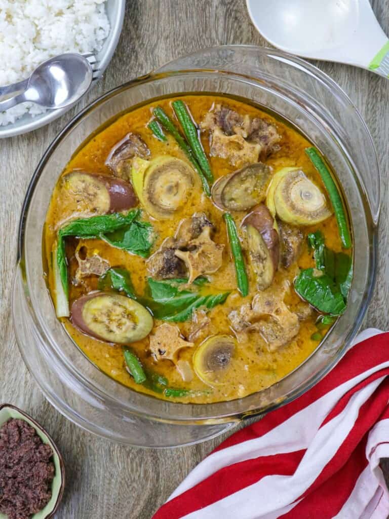 kare-kare in a glass serving bowl with a plate of steamed rice in the background.