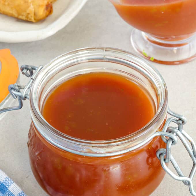 Sweet and Sour Sauce in a glass jar wtih fried lumpia in the background.