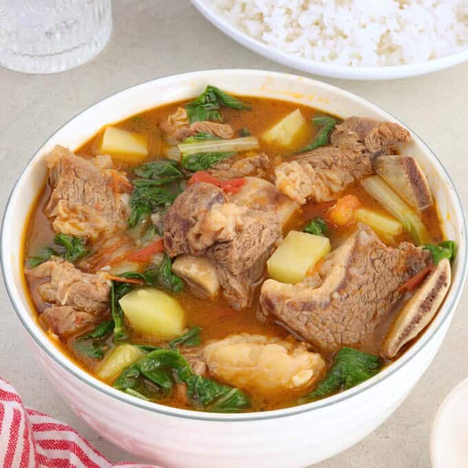 beef potato cabbage soup in a white bowl with a plate steamed rice and a glass of water in the background.