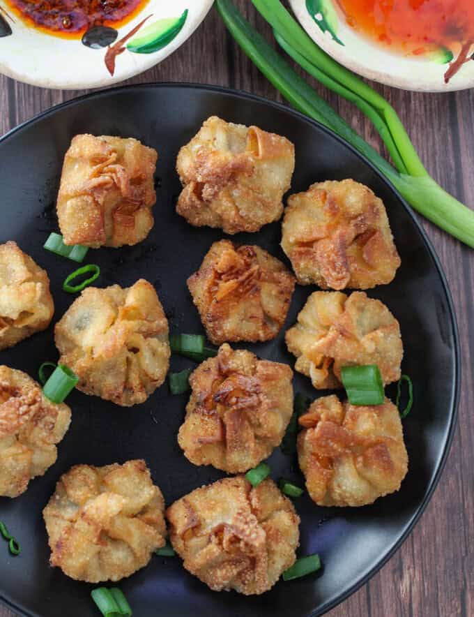 crispy fried longganisa wontons on a black plate with bowls of sweet chili sauce in the background.