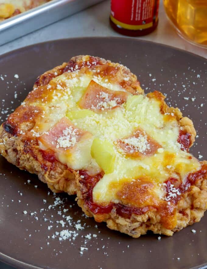 crispy breaded chicken breast baked with cheese, ham, and pineapple toppings on a brown plate.