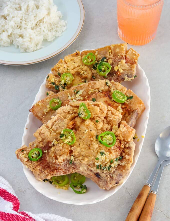 salt and pepper pork chops on a white platter with a plate of steamed rice in the background.