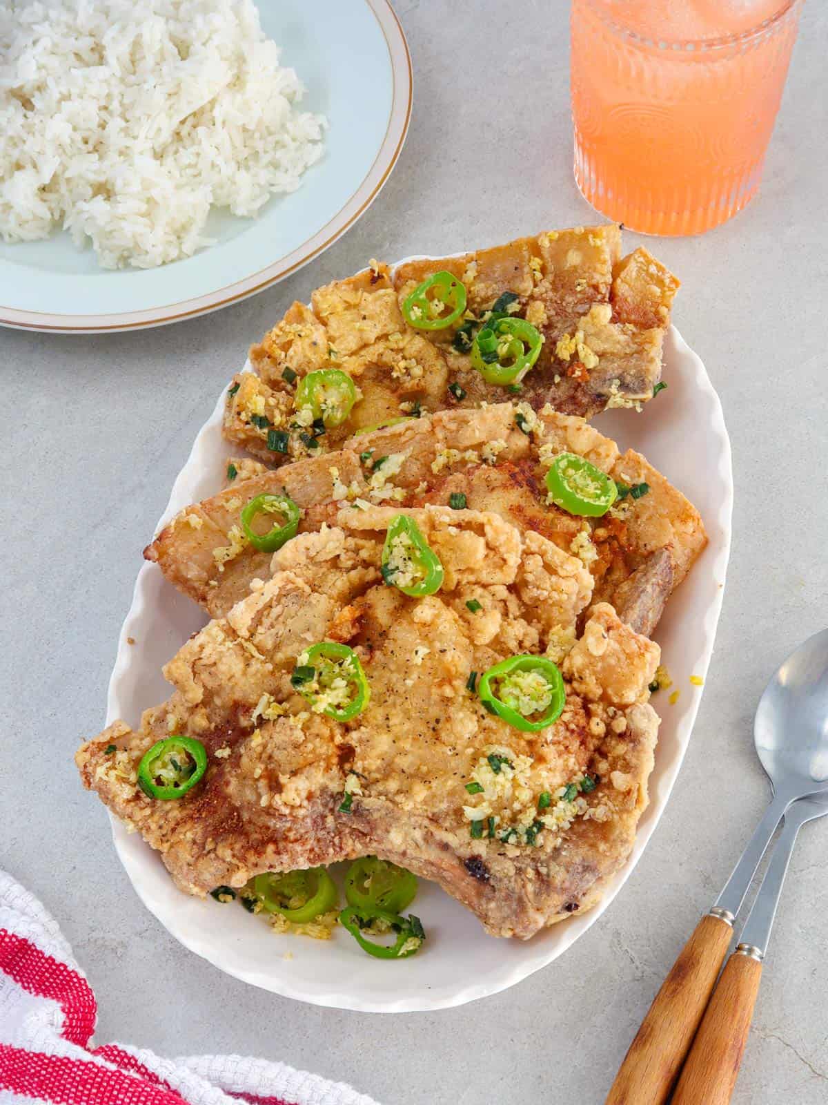 salt and pepper pork chops on a white platter with a plate of steamed rice in the background.