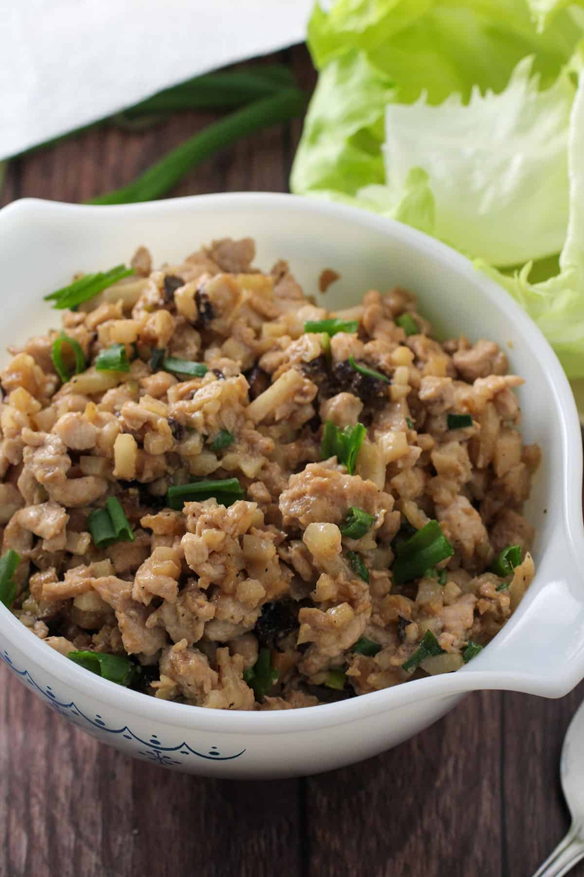 minced chicken cooked with green onions in a white bowl with iceberg lettuce leaves in the background.