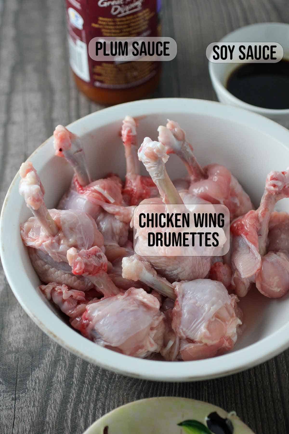 chicken drummettes formed into lollipops and soy sauce in individual bowls with bottle of plum sauce in the background.