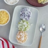 pichi-pichi with different flavors and coated with grated coconut on a white platter with a bowl of shredded cheese in the background.