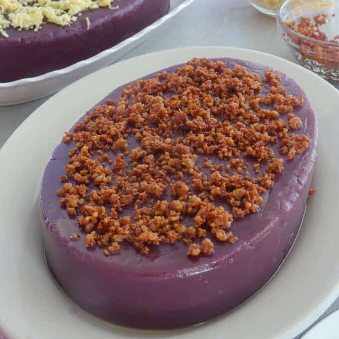 ube halaya topped with latik on a white serving platter with a ube halaya topped with cheese in the background.