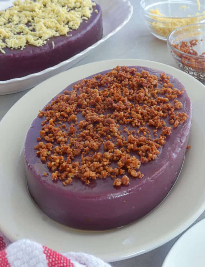 ube halaya topped with latik on a white serving platter with a ube halaya topped with cheese in the background.