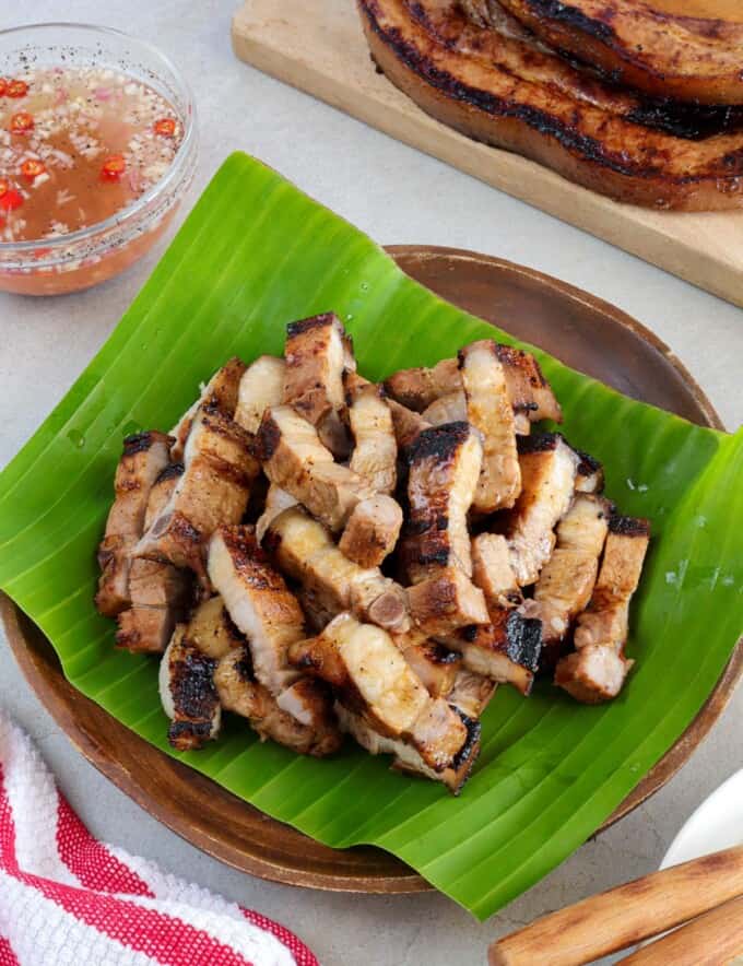 sliced Inihaw na Liempo on a banana leaf-lined wooden plate with a bowl of vinegar dipping sauce in the background.
