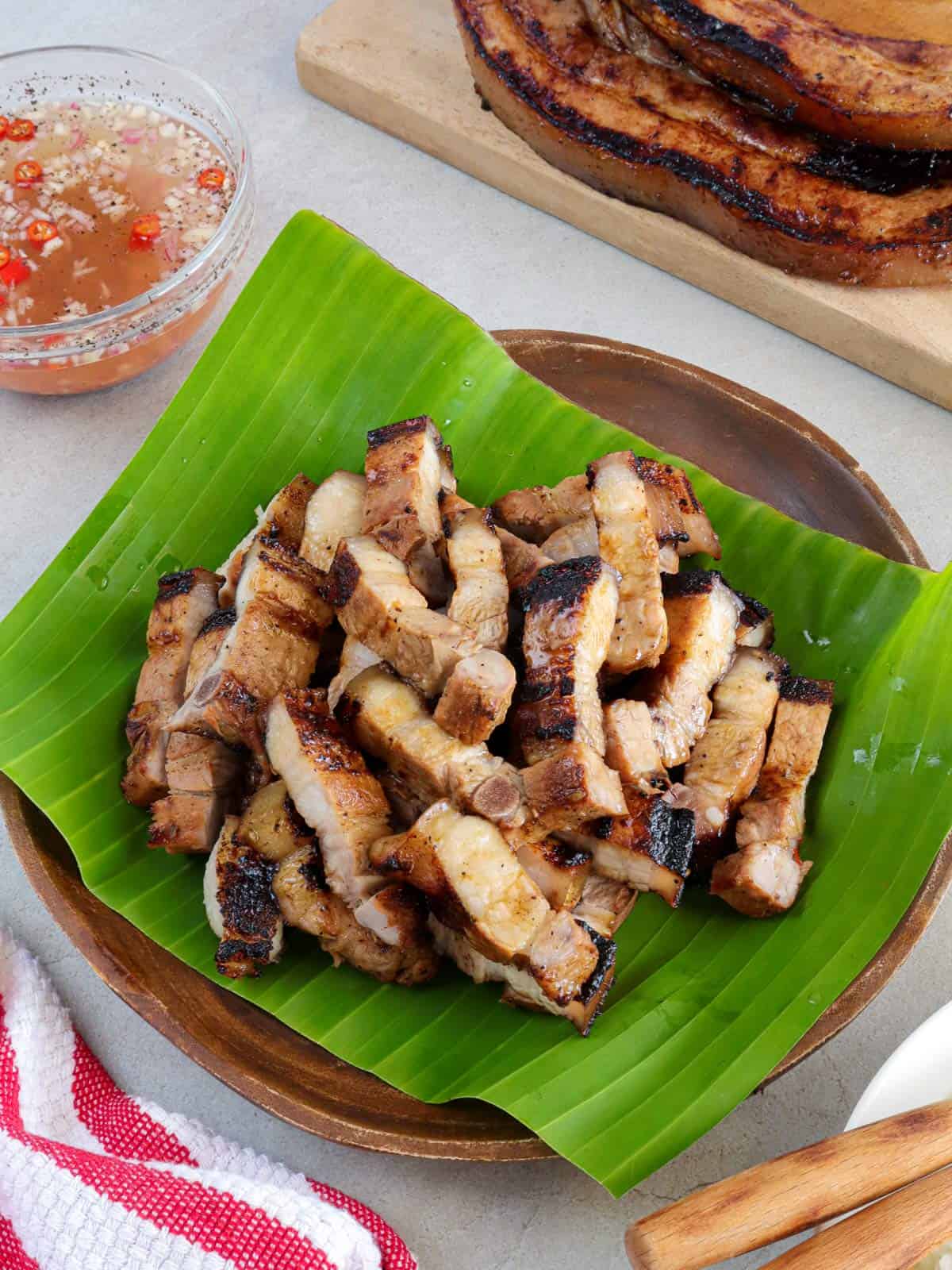 sliced Inihaw na Liempo on a banana leaf-lined wooden plate with a bowl of vinegar dipping sauce in the background.