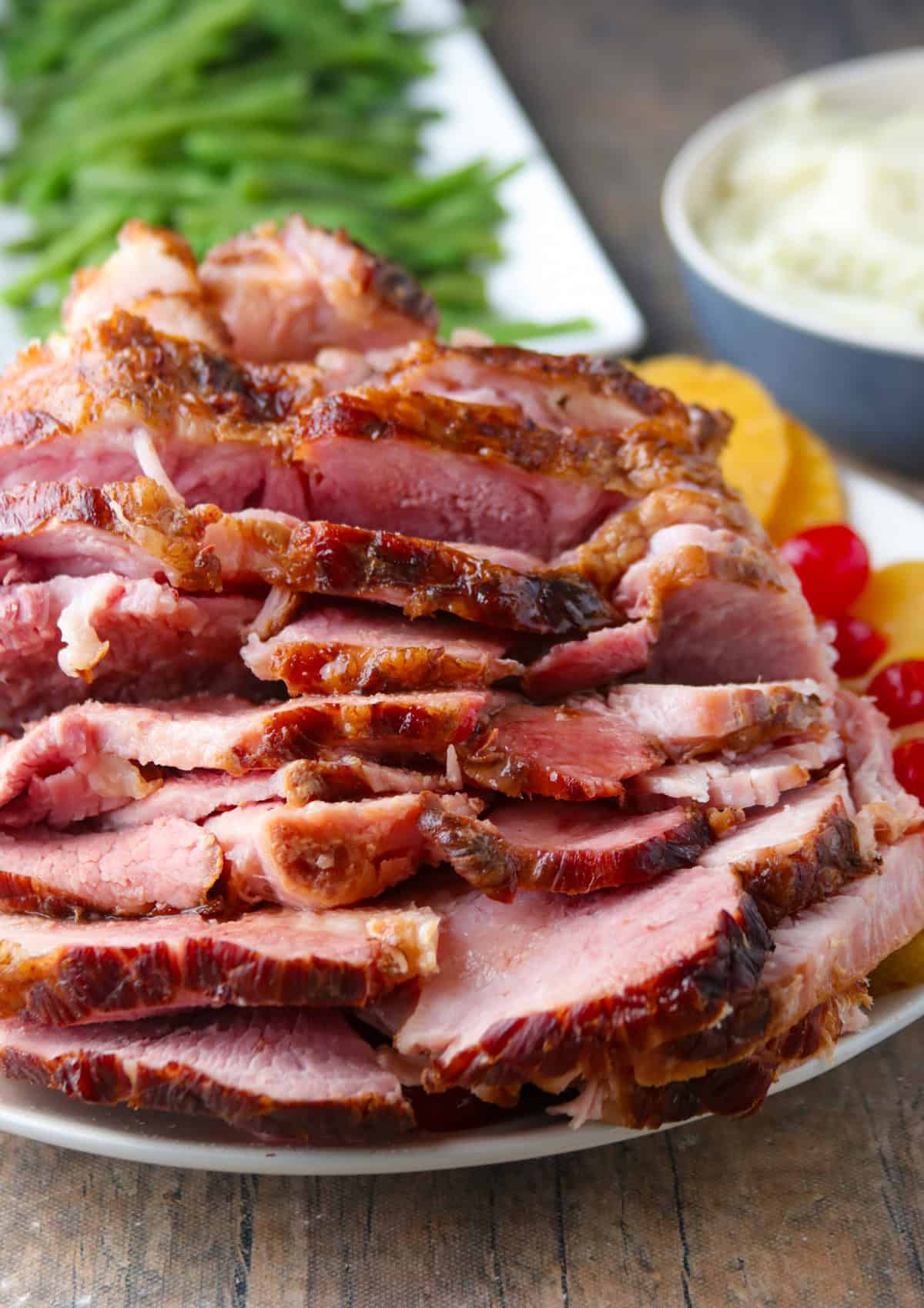 sliced ham, pineapple rings, and cheeries on a white platter with a plate of green beans in the background.