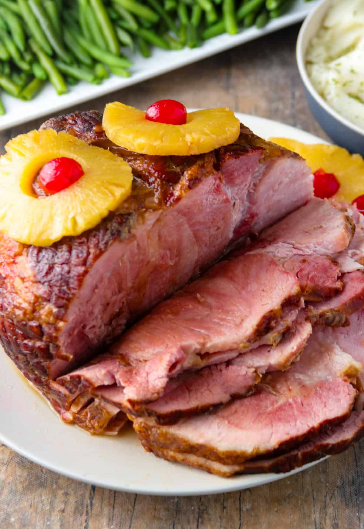 Slow Cooker Ham with Pineapples and maraschino cherries on a serving platter with a plate of green beans in the background.