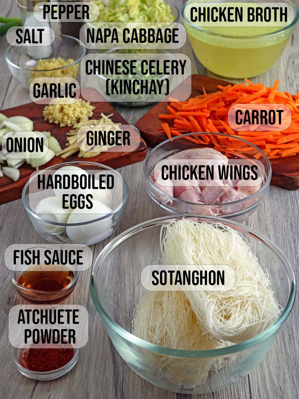 chicken wings, cellophane noodles, chopped onion, minced garlic, julienned carrots, hardboiled eggs, fish sauce, atsuete, powder in bowls and salt and pepper shakers in the background.