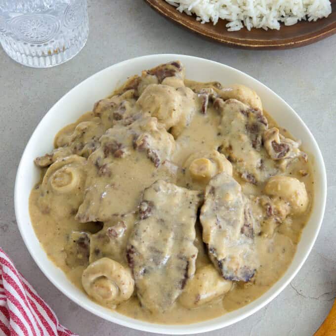 creamy beef and mushrooms in a white bowl with a plate of steamed rice in the background.