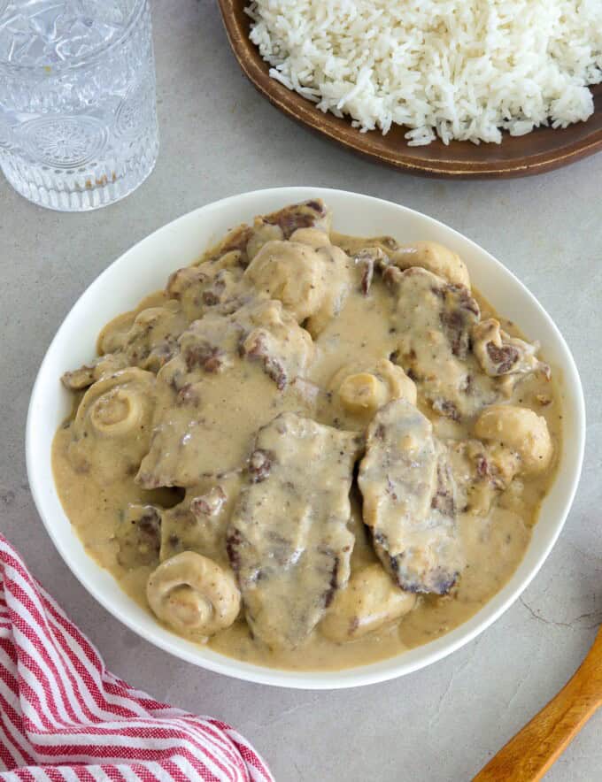 creamy beef and mushrooms in a white bowl with a plate of steamed rice in the background.