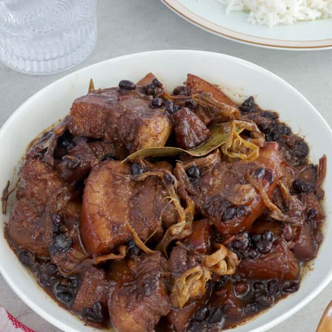 pork humba in a white serving bowl with a plate of steamed rice and glass of water in the background.