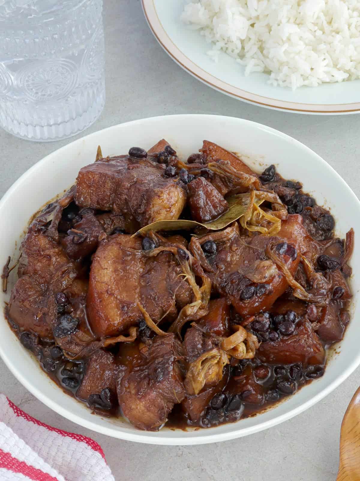 pork humba in a white serving bowl with a plate of steamed rice and glass of water in the background.