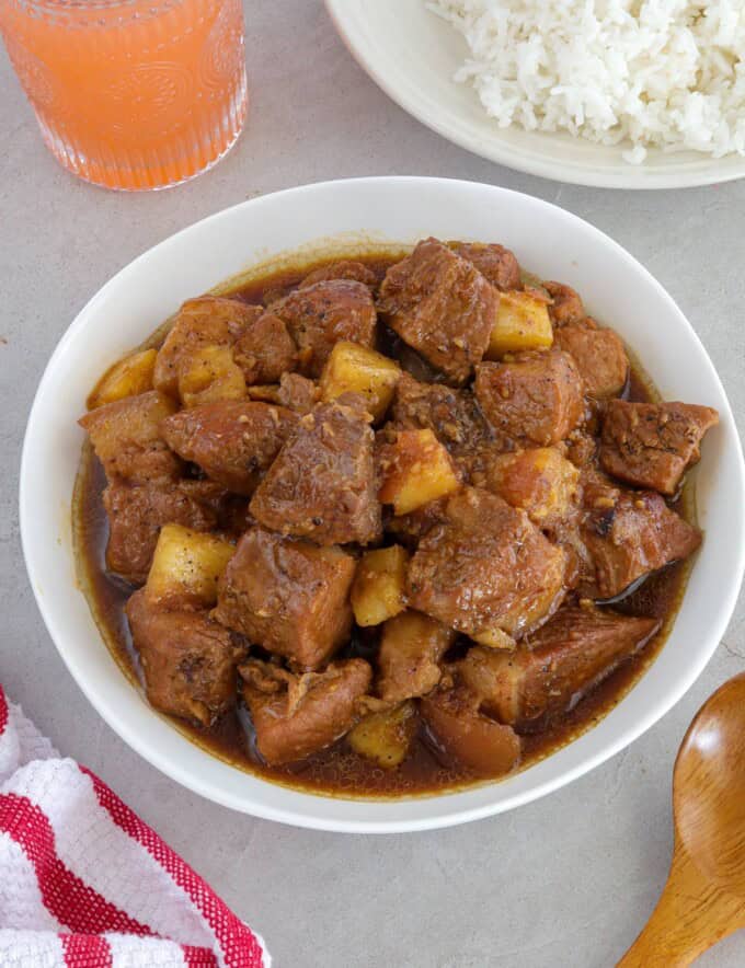 pork adobo with pineapple in a white serving bowl with a plate of steamed rice in the backround.