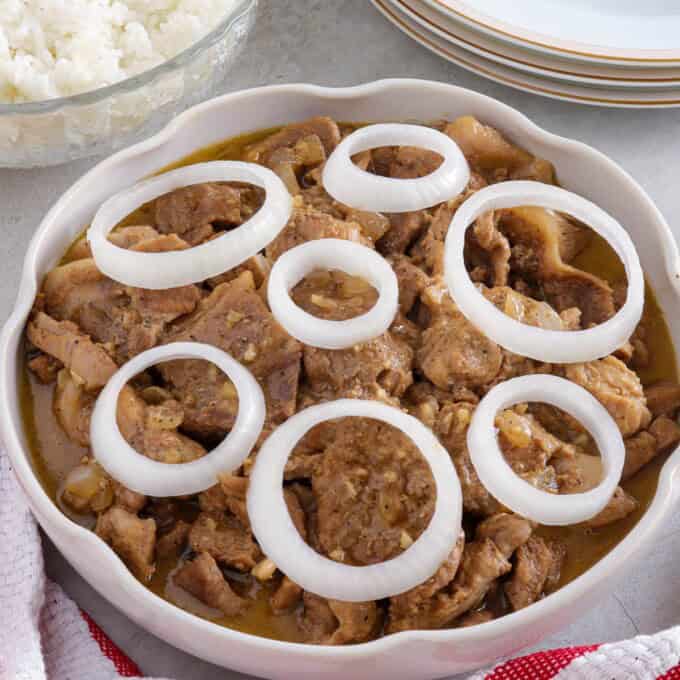 Creamy Pork Steak with onion rings in a white serving bowl with a bowl of steamed rice in the background.