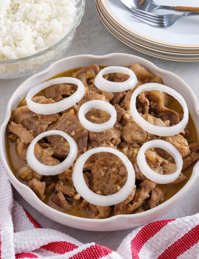 Creamy Pork Steak with onion rings in a white serving bowl with a bowl of steamed rice in the background.