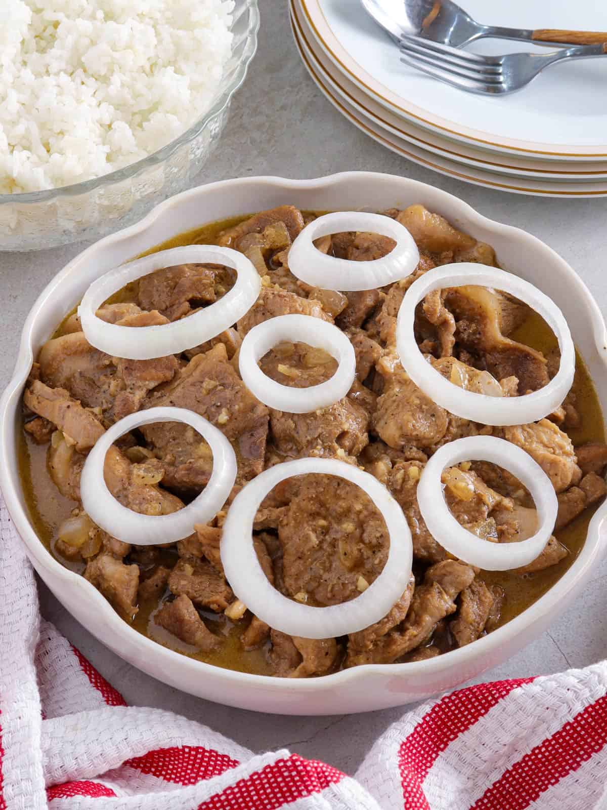 Creamy Pork Steak with onion rings in a white serving bowl with a bowl of steamed rice in the background.