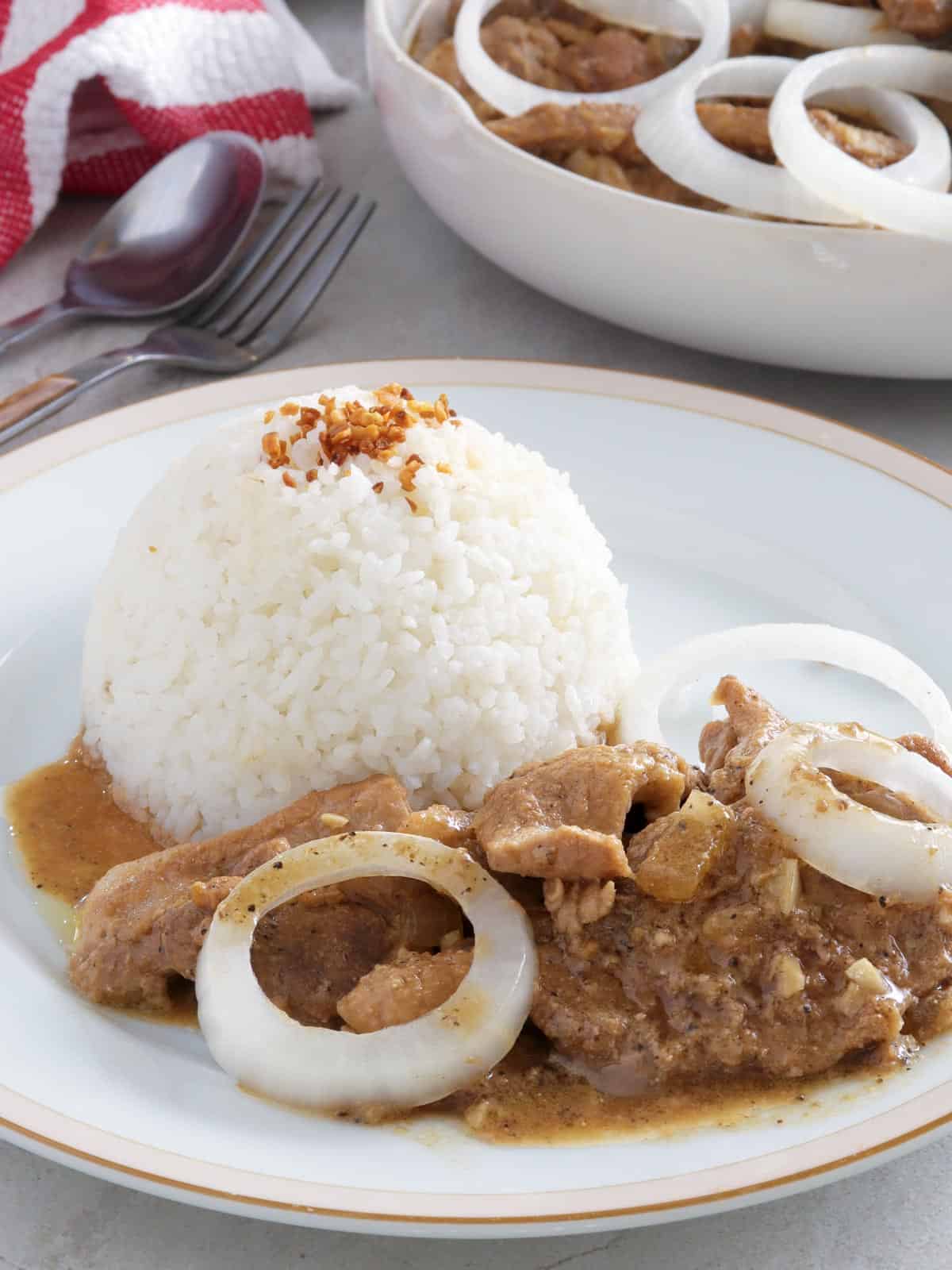 Creamy pork bistek on a white plate with steamed rice on the side.