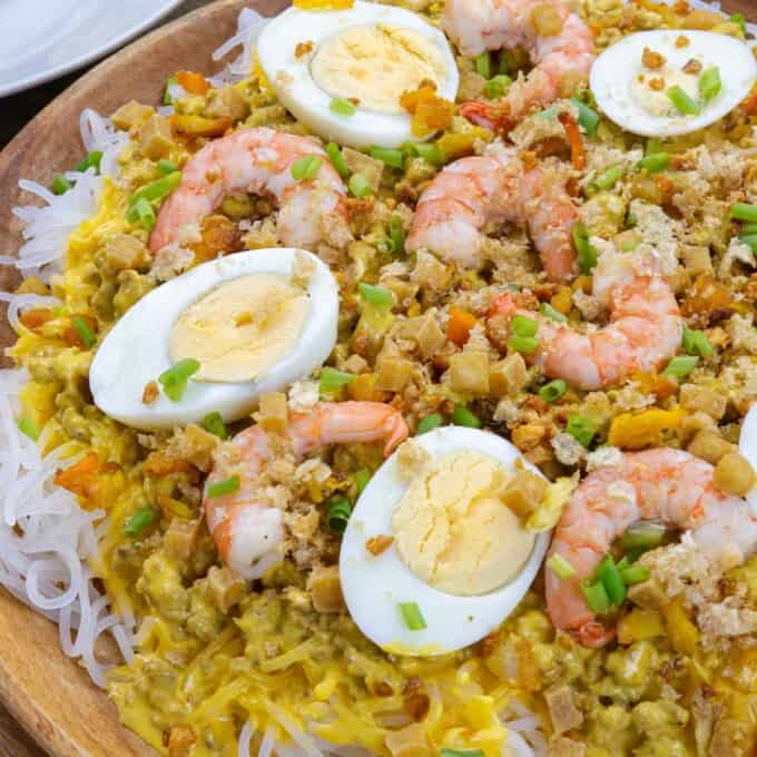 Creamy Palabok on a wooden platter with shrimp, hardboiled eggs, green onions, fried tofu toppings.