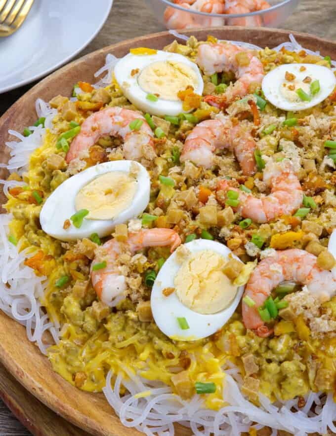 Creamy Palabok on a wooden platter with shrimp, hardboiled eggs, green onions, fried tofu toppings.